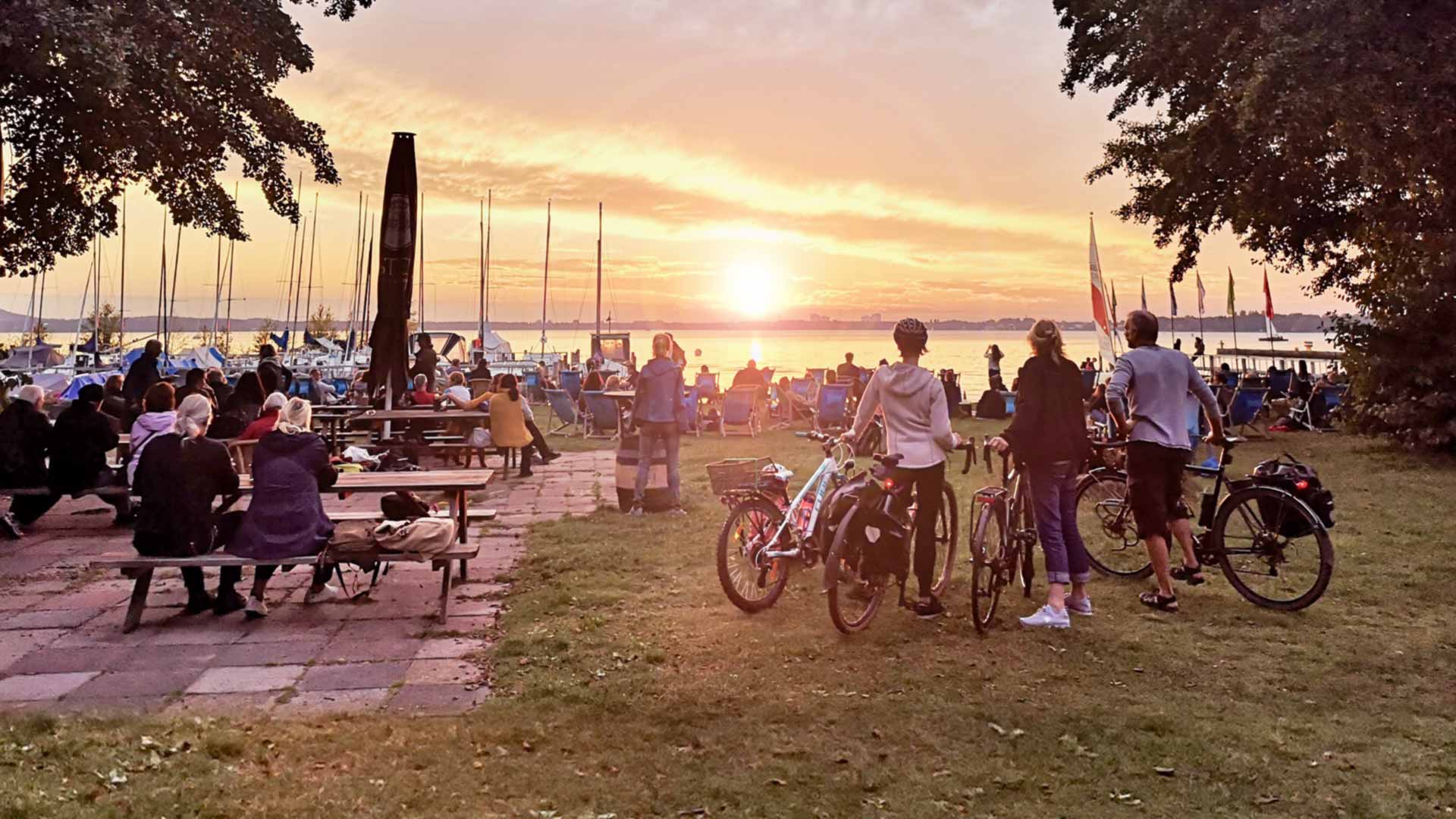 Gäste mit Fahrrädern genießen den Sonnenuntergang bei der Surf- und Segelschule am Müggelsee in Berlin-Köpenick